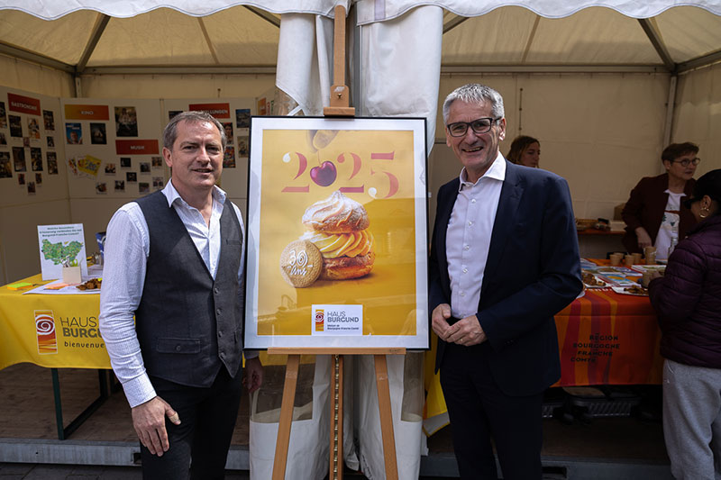 Landtagspräsident Hendrik Hering steht mit einer weiteren Person vor dem Stand. Zwischen ihnen befindet sich das gerahmte Plakat zum 30jährigen Bestehen des Hauses Burgund.