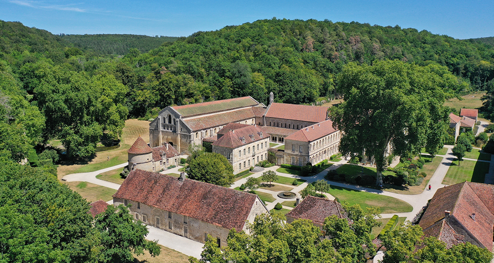 Auf dem Bild sieht man eine historische Klosteranlage in einer ländlichen, waldreichen Umgebung. Die Gebäude bestehen aus einer großen Kirche mit langem Dach und angrenzenden Klostergebäuden, die einen Innenhof umschließen. Die Anlage ist von gepflegten Gärten, Wegen und viel Grün umgeben, dahinter erstrecken sich dichte Wälder und sanfte Hügel. 