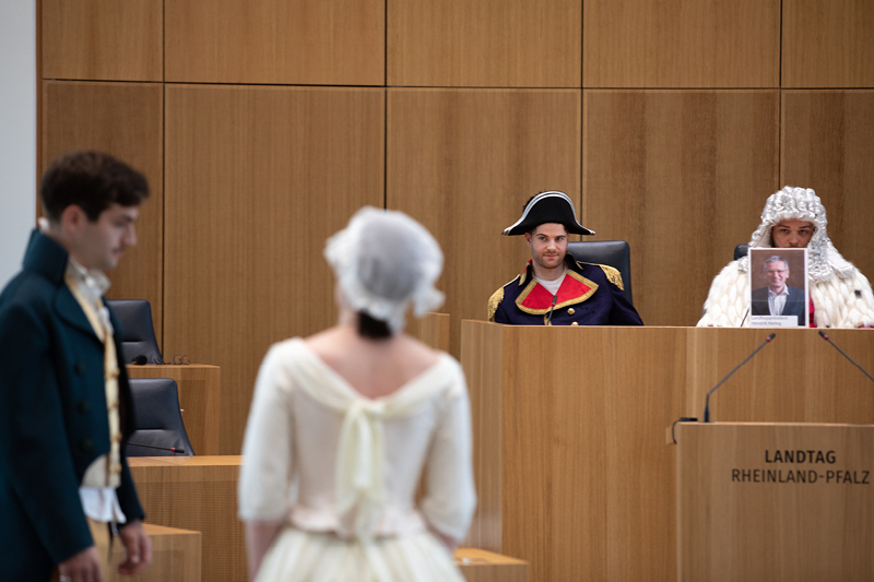 Auf dem Bild sieht man eine szenische Aufführung im Plenarsaal des Landtags Rheinland-Pfalz. Im Vordergrund stehen zwei Schauspieler in historischen Kostümen:  Der männliche Darsteller trägt einen grünen Gehrock mit goldenem Hemd und Weste.  Die weibliche Darstellerin ist in einem weißen Kleid mit Spitzenhaube gekleidet.  Im Hintergrund sitzen zwei weitere Darsteller am Rednerpult.