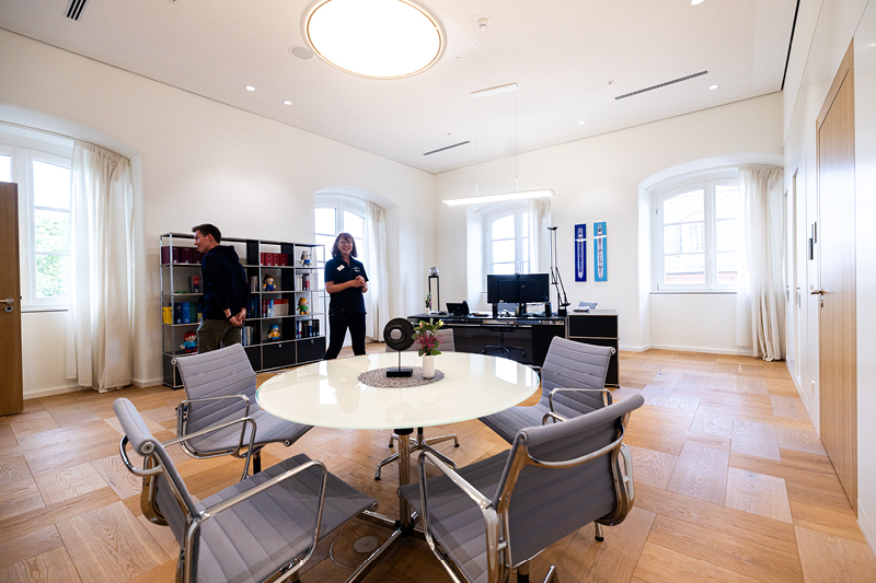 Auf dem Bild sieht man das Direktorenzimmer im Landtag Rheinland-Pfalz. Der Raum ist hell und freundlich gestaltet, mit großen Fenstern und eleganten Holzböden.  In der Mitte steht ein runder Besprechungstisch mit mehreren stilvollen Stühlen aus Metall und Stoff. Auf dem Tisch befindet sich eine kleine Blumenvase und ein dekorativer Untersetzer.  Im Hintergrund ist ein Schreibtisch mit zwei Monitoren zu sehen, daneben einige Dekorationsgegenstände und persönliche Elemente.  Links im Raum steht ein offenes Regal mit Büchern und bunten Objekten. An der Wand hängen zwei gerahmte Schilder/Banner.