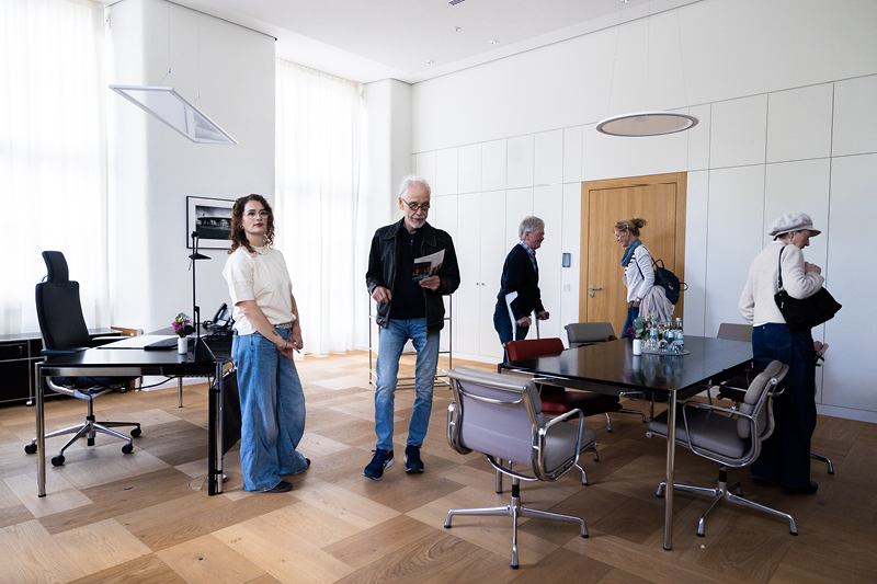 Auf dem Bild sieht man eine Gruppe von Personen im Präsidentenzimmer des Landtags Rheinland-Pfalz. Der Raum ist hell und großzügig gestaltet, mit großen Fenstern, die viel Tageslicht hereinlassen.  Auf der linken Seite steht ein eleganter Schreibtisch mit einem schwarzen Drehstuhl, einer Blumenvase und einem Telefon.  In der Mitte befindet sich ein großer Konferenztisch mit mehreren modernen Stühlen, auf dem Tisch stehen Wasserkaraffen und Gläser.  An der Wand sind große Schranktüren in einem minimalistischen Design zu sehen.