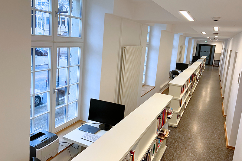 In einem langen Flur stehen mehrere aneinandergereihte Bücherregale. Neben ihnen befinden sich große Fenster und einzelne Tische mit Computermonitoren.