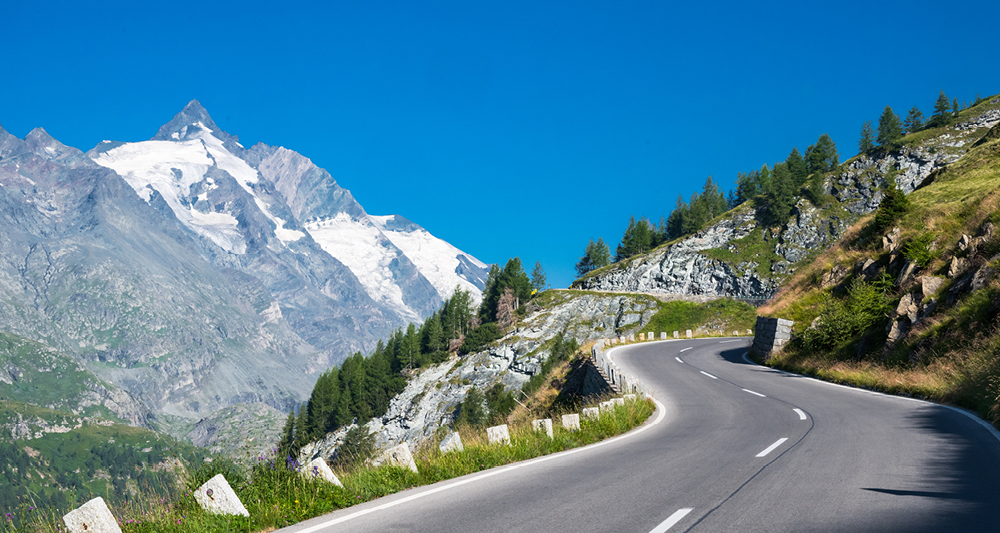 Zu sehen ist eine kurvenreiche Bergstraße in einer alpinen Landschaft. Die Straße windet sich am Hang entlang und ist von Felsen, Wiesen und Bäumen gesäumt. Im Hintergrund ragt ein hoher, schneebedeckter Berggipfel auf.