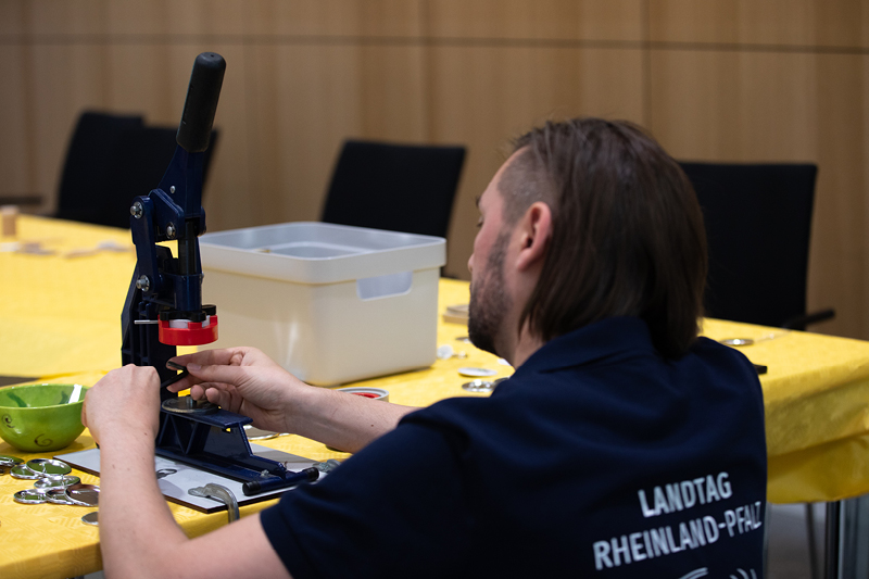Auf dem Bild sieht man einen Mann, der eine Buttonmaschine bedient. Der Mann trägt ein dunkelblaues T-Shirt mit der Aufschrift 
