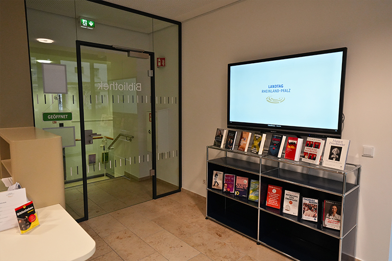 An einer Wand hängt ein Bildschirm mit der Aufschrift „Landtag Rheinland-Pfalz“ neben einer Glastür. Darunter steht ein Regal mit mehreren Büchern. Gegenüber befindet sich ein Tresen.