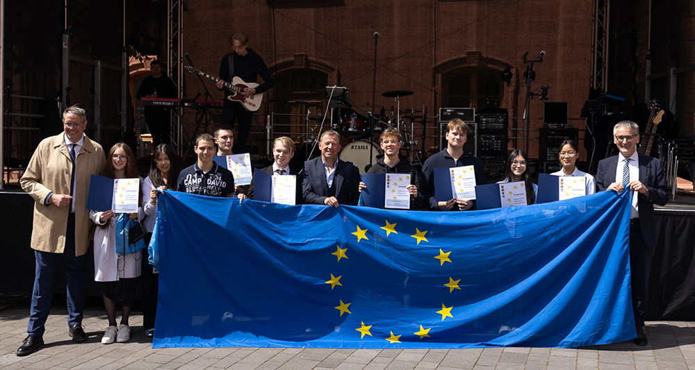 Eine Reihe von Menschen steht vor einer Bühne und hält gemeinsam eine Europaflagge. Einige Personen halten offene Urkundenmappen.
