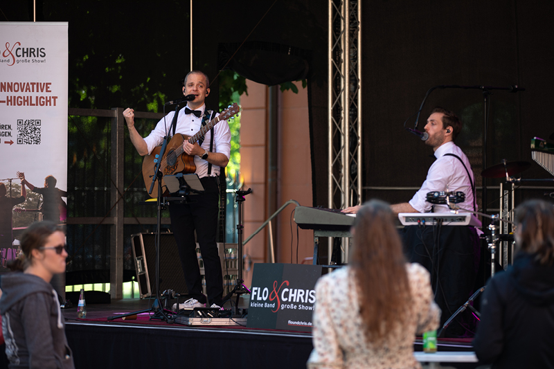 Auf dem Bild sieht man eine Live-Bühnenperformance des Duos Flo & Chris.  Bühne und Künstler Links auf der Bühne steht ein Musiker mit einer Gitarre, gekleidet in einem weißen Hemd mit schwarzer Fliege und Hosenträgern. Er hat ein Headset-Mikrofon und spricht oder singt ins Mikrofon.  Rechts sitzt ein weiterer Musiker an einem Keyboard, ebenfalls in einem weißen Hemd mit schwarzer Fliege und Hosenträgern, und singt in ein Mikrofon.