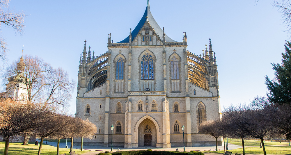Zu sehen ist ein majestätische Kirche. Um die Kirche sieht man eine grüne Landtschaft mit vielen Bäumen. Der Himmel ist strahlend Blau. Vor der Kirche befindet sich mittig eine Grünfläche.
