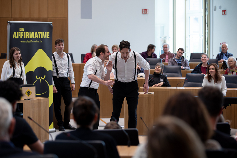 Auf dem Bild sieht man eine Gruppe von Schauspielern, die eine Improvisationstheater-Aufführung im Plenarsaal des Landtags Rheinland-Pfalz präsentieren. Die Schauspieler tragen weiße Hemden mit schwarzen Hosenträgern und schwarzen Hosen. Zwei männliche Schauspieler stehen im Vordergrund und agieren miteinander in einer lebhaften Szene, wobei einer dem anderen etwas ins Ohr flüstert.  Im Hintergrund stehen weitere Ensemblemitglieder, die das Geschehen beobachten und auf ihren Einsatz warten.  Links neben der Bühne steht ein großes Plakat mit der Aufschrift 