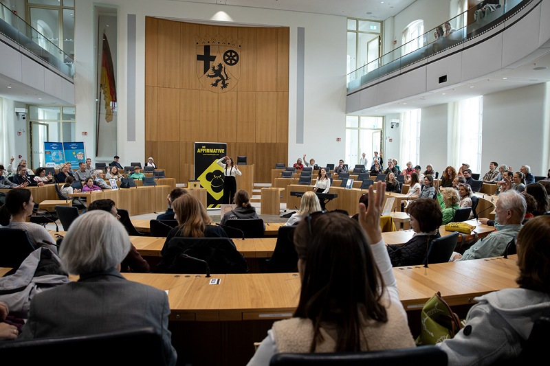 Zu sehen ist der Plenarsaal des Landtags Rheinland-Pfalz. Im Vordergrund stehen viele Menschen. Im Zentrum steht eine Person an einem Rednerpult. Hinter dieser Person ist ein auffälliges gelb-schwarzes Plakat mit der Aufschrift 