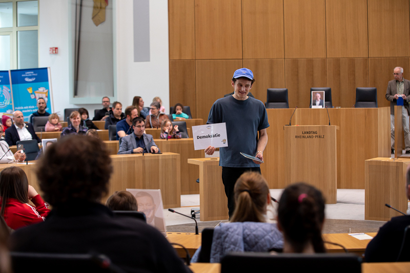 Auf dem Bild sieht man eine Veranstaltung im Plenarsaal des Landtags Rheinland-Pfalz. Im Vordergrund steht ein Mann mit einer blauen Kappe und einem dunkelblauen T-Shirt. Er hält ein Schild mit der Aufschrift 