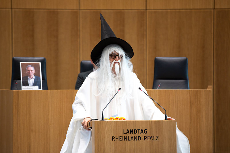 Auf dem Bild sieht man eine Person, die als Zauberer verkleidet ist. Die Person trägt ein langes weißes Gewand, eine weiße Perücke mit langem Haar und Bart sowie einen großen schwarzen Spitzhut. Sie steht hinter dem Rednerpult im Plenarsaal des Landtags Rheinland-Pfalz.