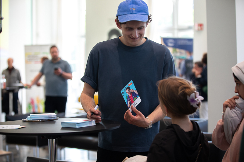 Auf dem Bild sieht man einen jungen Mann mit einer blauen Kappe und einem dunkelblauen T-Shirt. Er steht an einem Stehtisch und signiert Autogrammkarten für Kinder. Der Mann lächelt freundlich und hält eine Karte mit seinem Bildmotiv in der Hand.