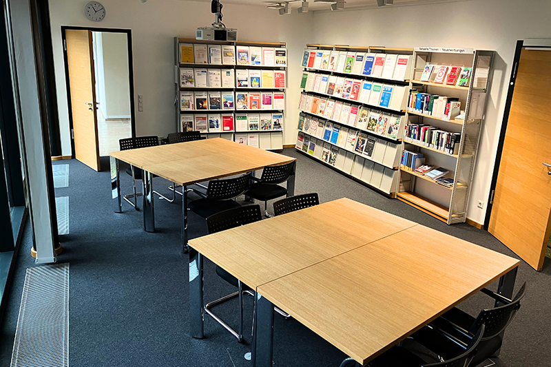 In der Mitte eines Raums stehen zwei rechteckige Holztische mit mehreren Stühlen. Im Hintergrund befinden sich Wandregale mit Zeitschriften und Büchern.