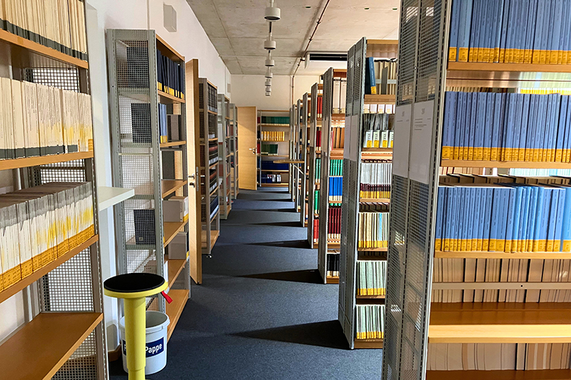 Ein breiter Gang führt zwischen mehreren Reihen von Bücherregalen hindurch.