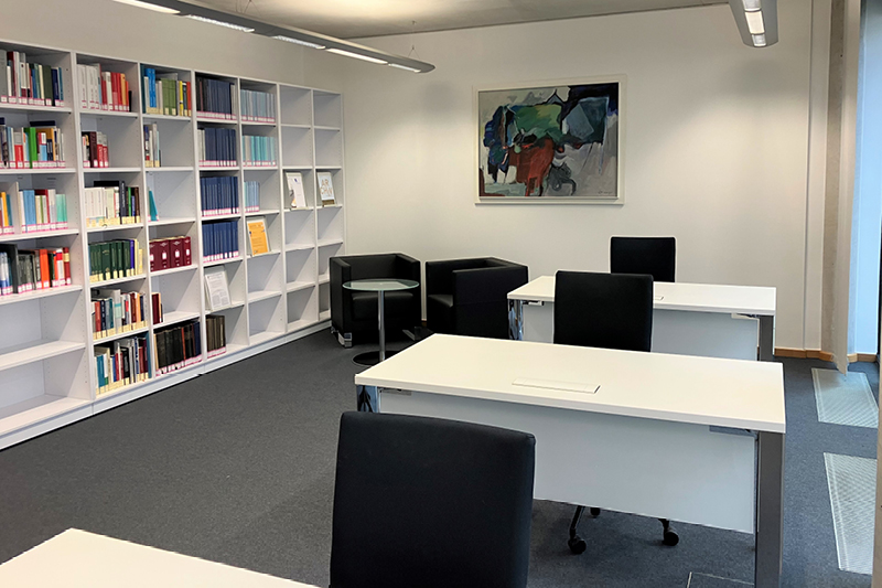 Zu sehen ist der Lesesaal des Landtagsarchivs im 1. OG. In dem Raum befinden sich mehrere weiße Tische mit schwarzen Schreibtischstühlen. Auf der linken Seite befindet sich ein großes Bücherregal. Rechts davon hängt ein Gemälde.