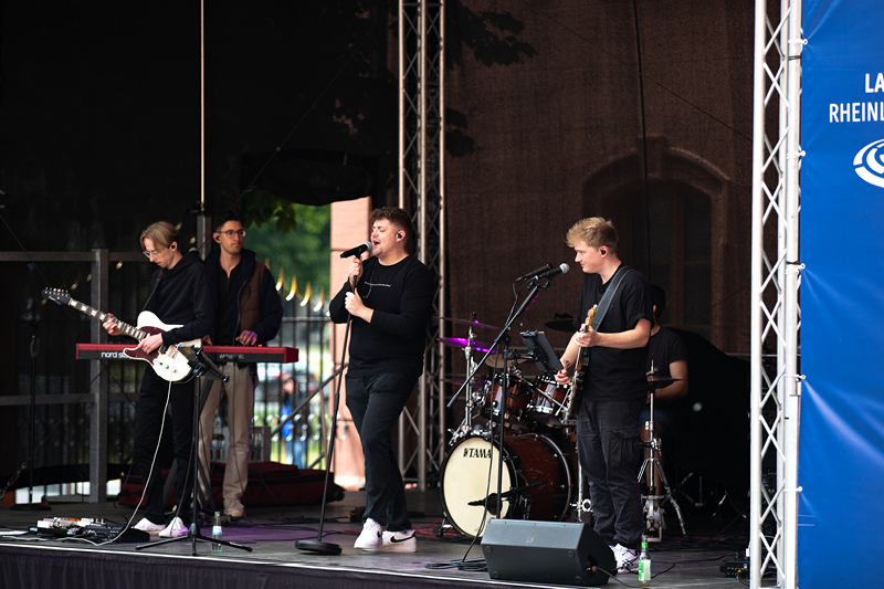 Auf dem Bild sieht man eine Live-Band, die auf einer Bühne während des Verfassungsfests 2025 im Freien auftritt. Die Band besteht aus mehreren jungen Musikern, die schwarze Kleidung tragen.  Im Vordergrund steht der Leadsänger mit einem Mikrofon, während ein Gitarrist rechts daneben ebenfalls in das Mikrofon singt.  Links auf der Bühne steht ein weiterer Gitarrist, daneben ein Keyboarder.  Im Hintergrund sitzt ein Schlagzeuger hinter einem Yamaha-Schlagzeug und begleitet den Auftritt rhythmisch.
