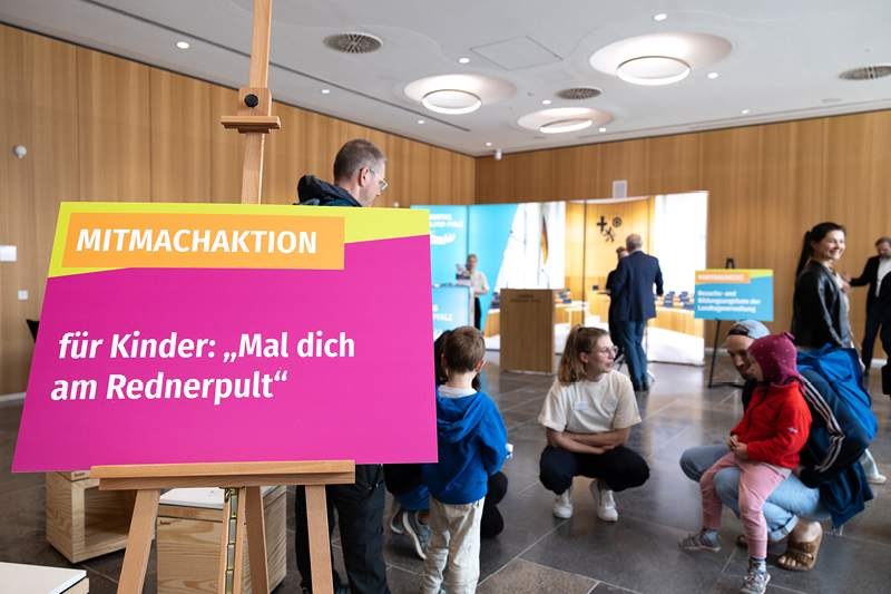 Auf dem Bild sieht man eine Mitmachaktion für Kinder im Rahmen einer Veranstaltung im Landtag Rheinland-Pfalz. Im Vordergrund steht ein buntes Schild auf einer Staffelei mit der Aufschrift: 