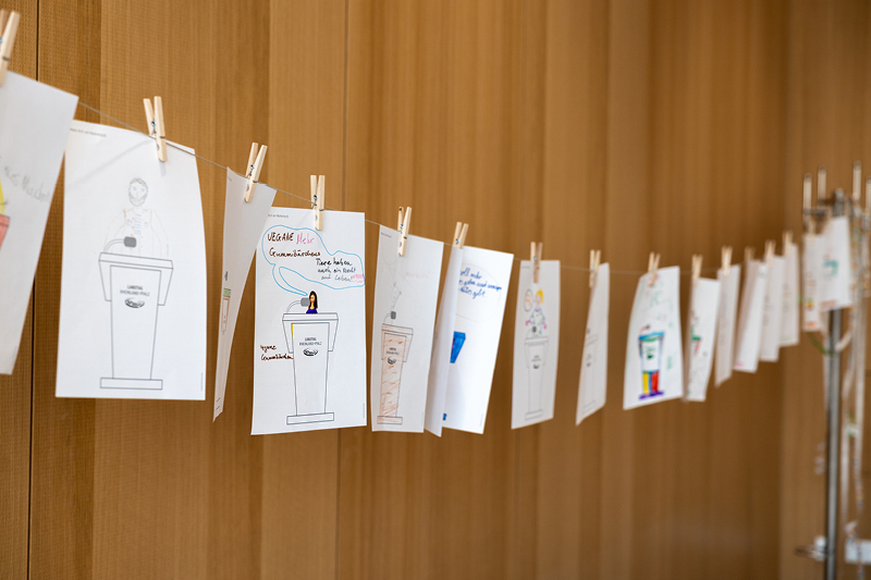 Auf dem Bild sieht man die Ergebnisse einer Mitmachaktion für Kinder im Rahmen einer Veranstaltung im Landtag Rheinland-Pfalz. Die Bilder sind auf einem dünnen Seil aufgehängt und mit kleinen Holzklammern befestigt.  Jedes Bild zeigt eine Zeichnung eines Rednerpults des Landtags Rheinland-Pfalz mit individuellen Ausgestaltungen.  Manche Zeichnungen sind farbig gestaltet, andere enthalten Sprechblasen oder kurze Botschaften.