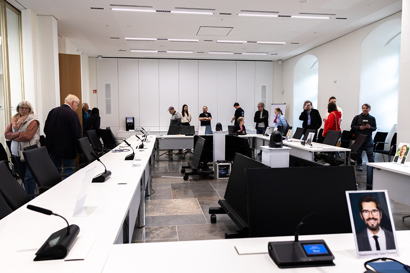 Auf dem Bild sieht man einen Sitzungssaal im Landtag Rheinland-Pfalz. Der Raum ist hell und modern gestaltet, mit großen Fenstern und weißen Wänden.  Die Sitzanordnung ist in U-Form angeordnet. Auf den Tischen stehen Mikrofone und Namensschilder, die die Positionen der Teilnehmer markieren.  Vor einigen Plätzen sind Porträtfotos aufgestellt, die die Position von bestimmten Abgeordneten darstellen.