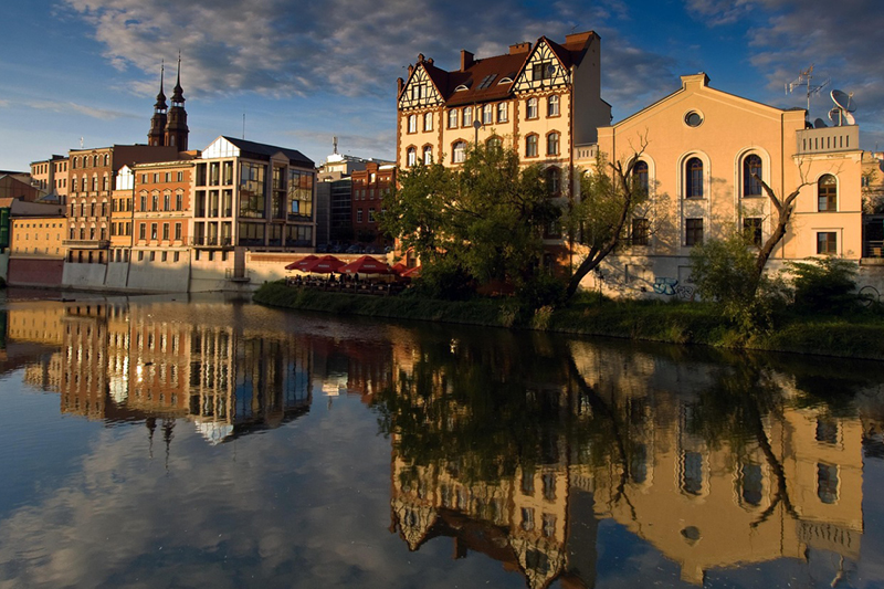 Auf dem Bild ist eine malerische Uferansicht einer Stadt zu sehen. Die Gebäude spiegeln sich klar in einem ruhigen Fluss. Entlang des Wassers sind Bäume und Sträucher zu sehen. Ganz links ragen zwei Kirchtürme in den Himmel.