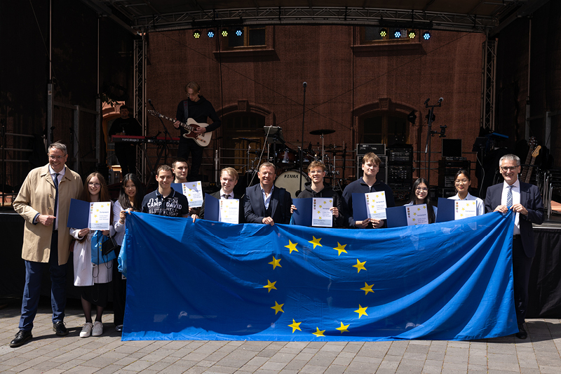 Auf dem Bild sieht man eine Gruppe von Personen, die vor einer Bühne stehen und eine große Europaflagge mit gelben Sternen auf blauem Hintergrund präsentieren.