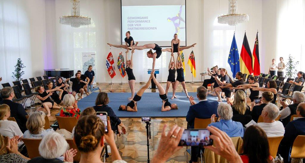 Akrobat:innen bilden in der Mitte eines Saals eine Pyramide. Das Publikum sitzt auf Stühlen um sie herum. Im Hintergrund befinden sich verschiedene Flaggen und eine Leinwand.