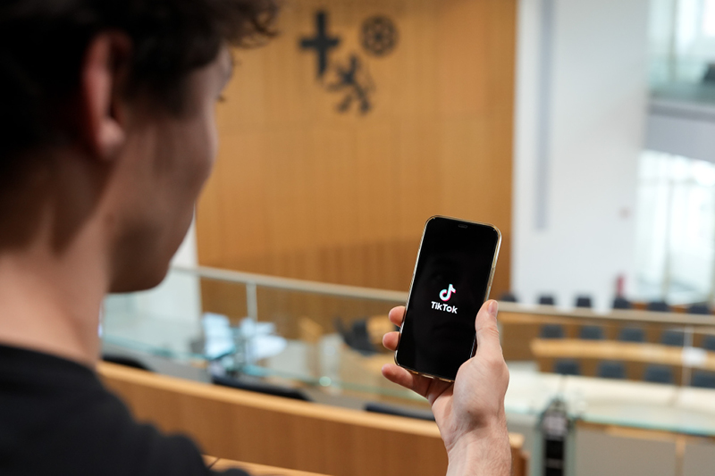 Zu sehen ist ein junger Mann. Er steht auf der Besuchertribüne des Plenarsaals und hält ein Smartphone in der Hand. Auf dem Bildschirm sieht man das TikTok Logo.