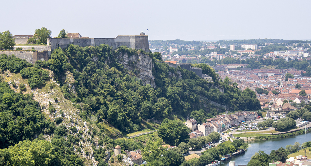 Eine massive Festung thront auf einem bewaldeten Hügel. Am Fuß des Hügels liegt
eine Stadt, durch die ein Fluss fließt.