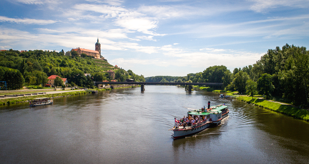 Zu sehen ist ein Fluss. Auf dem Fluss schwimmt ein kleines Boot. Auf diesem befinden sich viele Menschen. Seitlich des Flusses sieht man eine grüne Landschaft mit einer Kapelle. Der Himmel leuchtet blau.