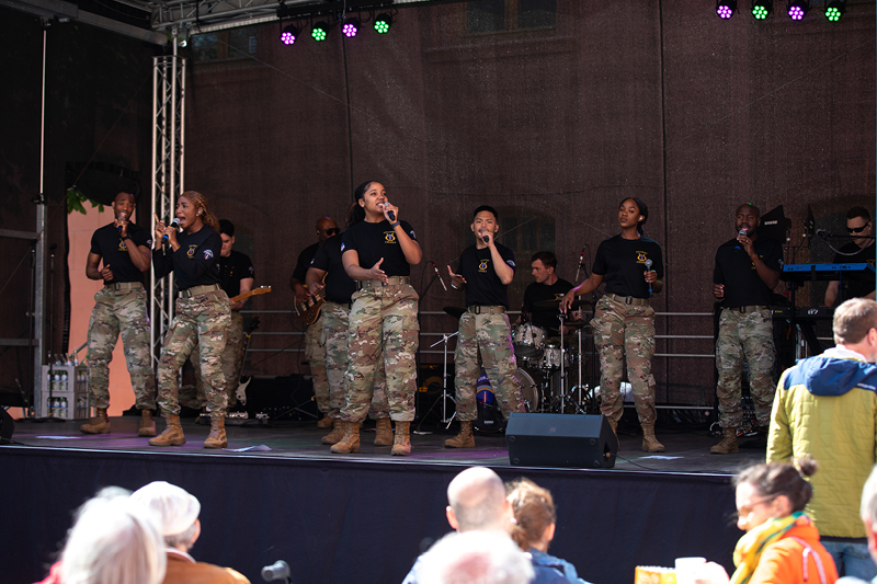 Auf dem Bild sieht man eine musikalische Aufführung auf einer Bühne im Freien. Die Gruppe besteht aus mehreren Sängerinnen und Sängern sowie Musikern, die Militäruniformen tragen. Es handelt sich um die USAREUR-AF Band and Chorus. Die Künstler:innen tragen schwarze T-Shirts mit einem Emblem und militärische Tarnhosen sowie Stiefel.  Die Frontsängerinnen und -sänger singen in Mikrofone, während die Band im Hintergrund spielt. Zu sehen sind unter anderem ein Schlagzeuger, ein Gitarrist und ein Keyboarder.