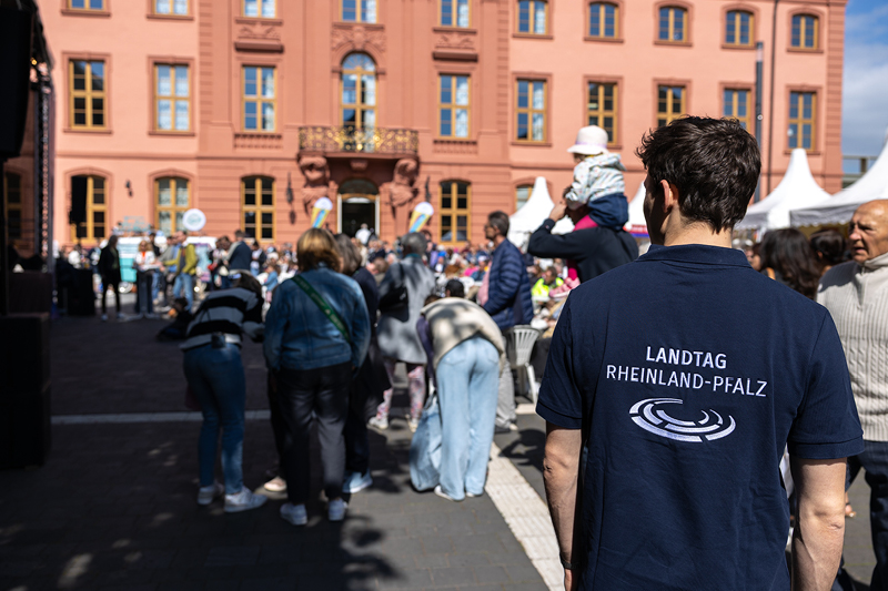 Im Vordergrund steht ein Mann mit einem dunkelblauen T-Shirt, auf dessen Rückseite die Aufschrift 