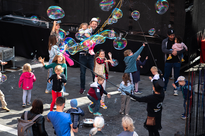 Auf dem Bild sieht man eine fröhliche Szene während des Verfassungsfests, bei der Kinder und Erwachsene mit großen Seifenblasen spielen. Im Mittelpunkt stehen Kinder, die begeistert nach großen Seifenblasen greifen und versuchen, diese zu fangen oder platzen zu lassen.  Eine Person, die zum Event-Team zu gehören scheint, produziert die Seifenblasen mit langen Stäben und Seifenlauge.  Erwachsene begleiten die Kinder und beobachten das Geschehen, einige halten Kleinkinder auf dem Arm.