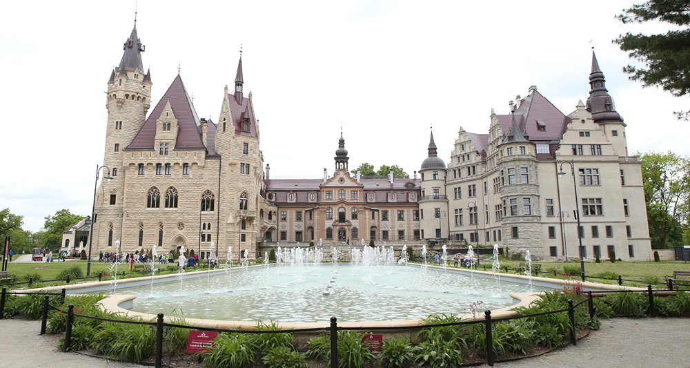 Auf dem Bild ist das Schloss Moszna (polnisch: Zamek w Mosznej) in Polen zu sehen. Es ist ein prachtvolles Schloss mit einer Mischung aus Barock-, Neorenaissance- und neugotischen Baustilen. Im Vordergrund befindet sich ein Springbrunnen, der Teil der gepflegten Schlossanlage ist. Das Schloss ist bekannt für seine zahlreichen Türme und romantische Architektur.