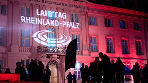 Zu sehen ist die Front des Deutschhauses bei Nacht. Die Hausfront wird mit buntem Licht angestrahlt, oberhalb des Eingangs wird das Logo auf die Hauswand projeziert. Im Vordergund sieht man einige Menschen stehen.