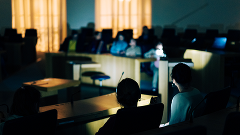 Zu sehen ist ein Teil des dunklen Plenarsaals. Man sieht die Umrisse einiger sitzenden Menschen.