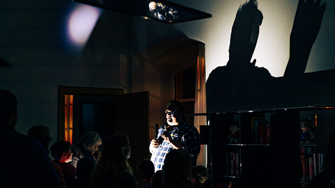 Zu sehen ist ein dunkler Raum. Mittig im Bild steht ein kräftiger Mann, der mit einer Taschenlampe angeleuchtet wird. Sein Schatten wird in Übergröße an die Wand geworfen. Man sieht die Umrisse einiger Kinder.