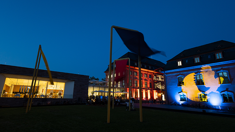 Zu sehen sind mehrere Gebäude bei Nacht, die angeleuchtet werden. Auf der rechten Seite sieht man das blau-gelbe Logo der Museumsnacht.
