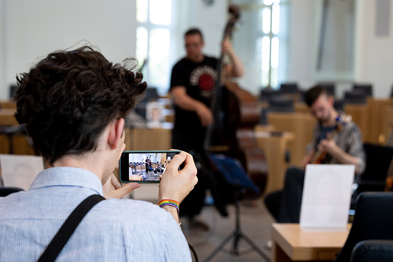 Zu sehen ist ein Besucher, der mit seinem Smartphone Musiker im Hintergrund aufnimmt.
