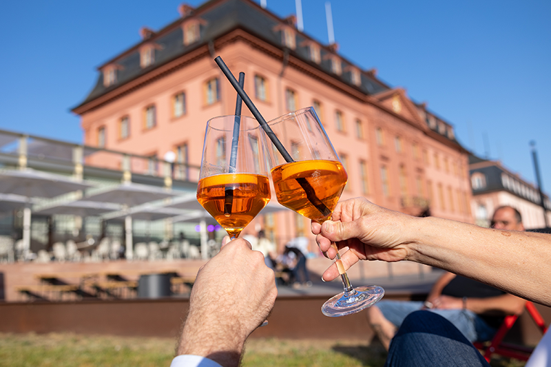 Zu sehen sind zwei Hände mit Aperol gefüllten Gläsern, die sich zu prosten. Im Hintergrund das Deutschhaus mit Restaurant. 