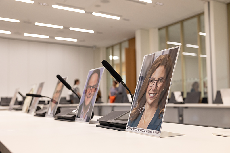 Zu sehen sind nebeneinder stehende Tische, auf denen Fotos von einigen Abgeordenten des Landtags stehen sowie dazwischenstehende Tischmikrophone.
