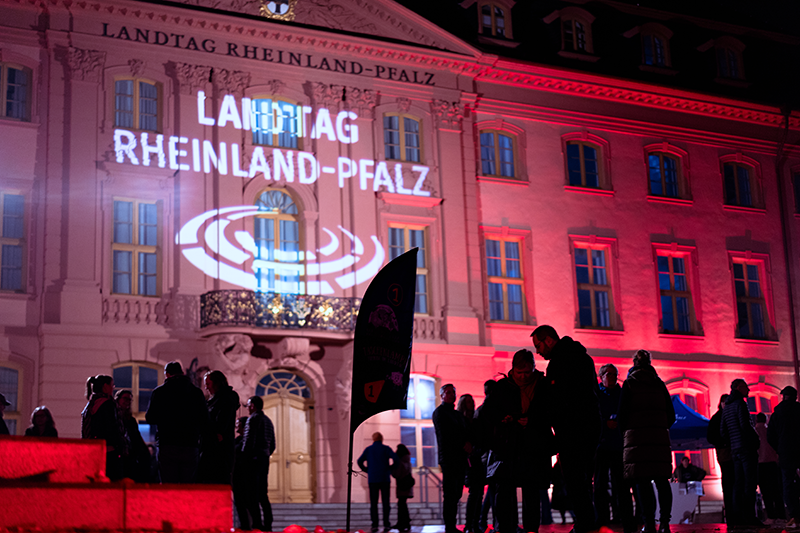 Zu sehen ist die Front des Deutschhauses bei Nacht. Die Hausfront wird mit buntem Licht angestrahlt, oberhalb des Eingangs wird das Logo auf die Hauswand projeziert. Im Vordergund sieht man einige Menschen stehen.