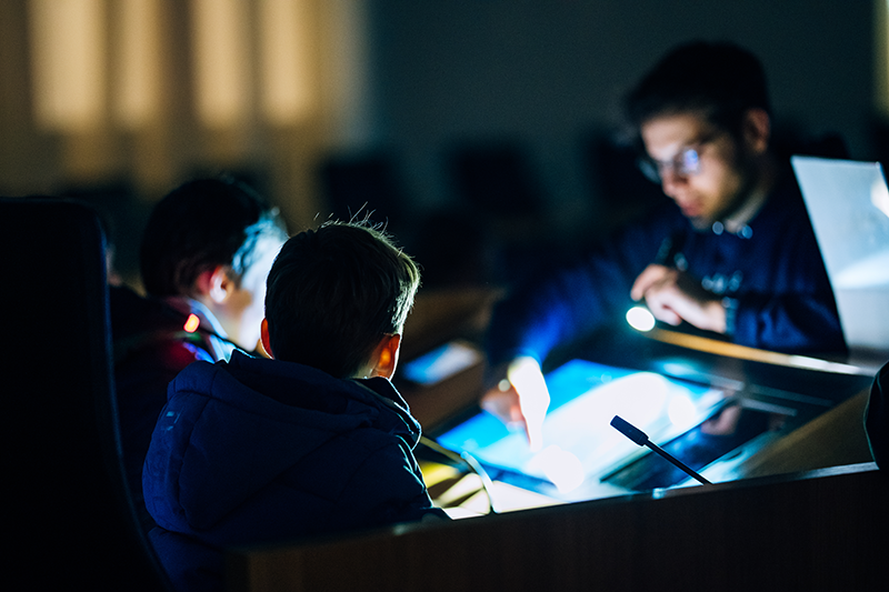 Zu sehen ist das Pult des Präsidenten im Plenarsaal im Dunkeln. Vor dem beleuchteten Pult stehen drei Kinder.