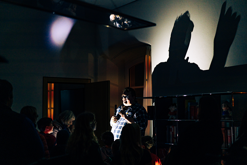 Zu sehen ist ein dunkler Raum. Mittig im Bild steht ein kräftiger Mann, der mit einer Taschenlampe angeleuchtet wird. Sein Schatten wird in Übergröße an die Wand geworfen. Man sieht die Umrisse einiger Kinder.