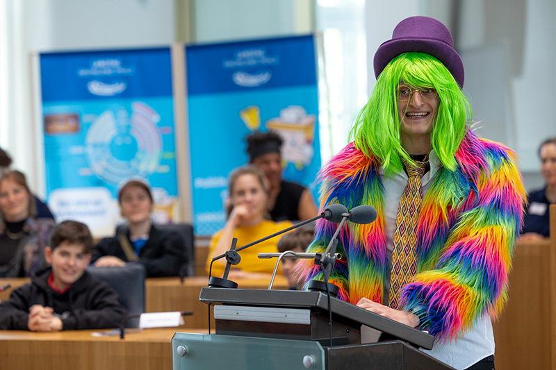 Checker Julian während seinem Auftritt im Plenarsaal. Er trägt bunte Klamotten und eine bunte Perrücke.