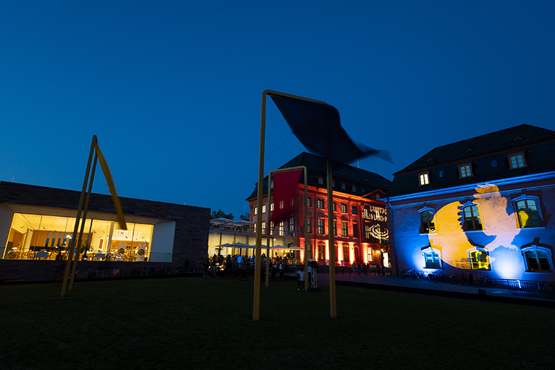 Zu sehen sind mehrere Gebäude bei Nacht, die angeleuchtet werden. Auf der rechten Seite sieht man das blau-gelbe Logo der Museumsnacht.